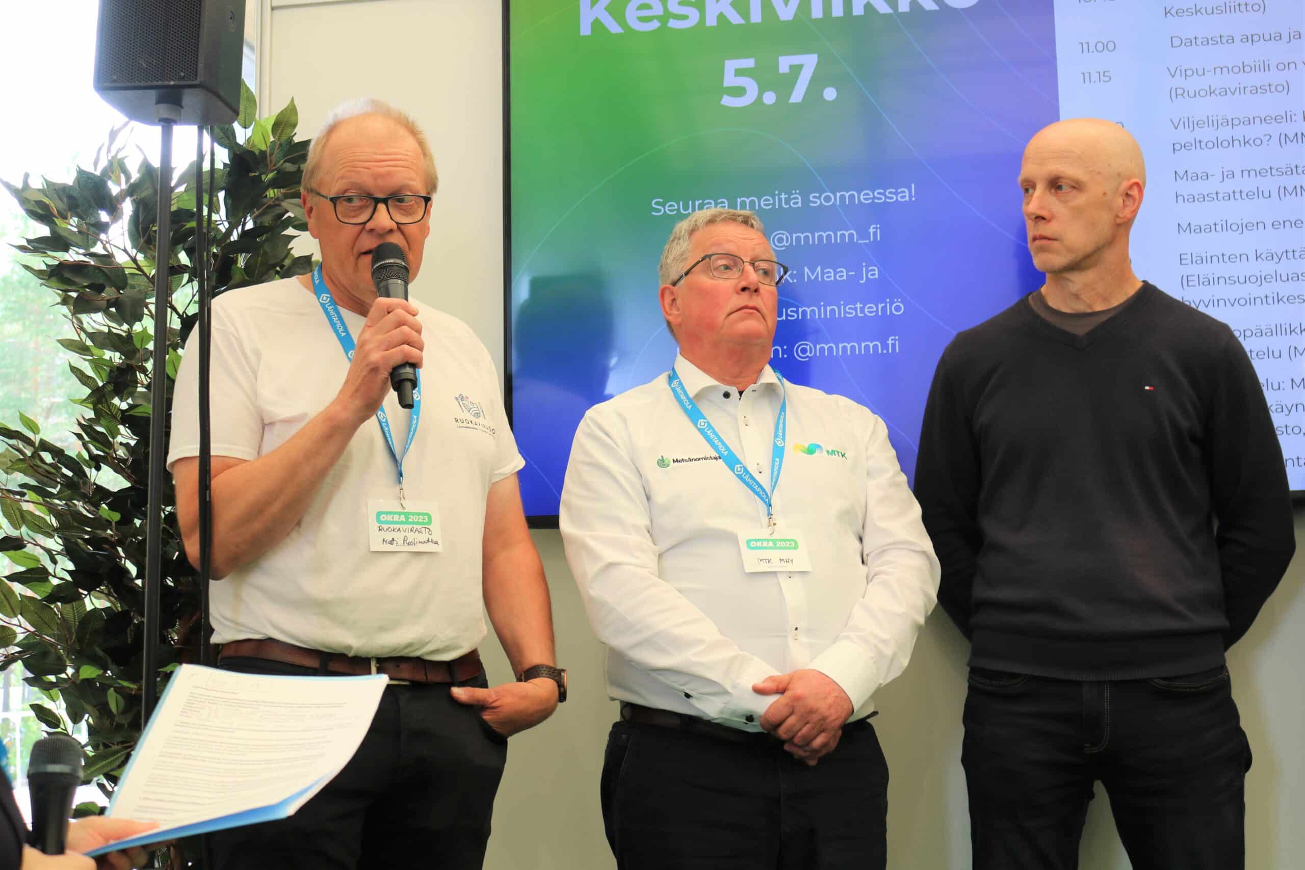 Kolme ihmistä lavalla. Vasemmalla mikrofonin kanssa Matti Puolimatka, keskellä Paavo Myllymäki ja oikealla Markku Paija.