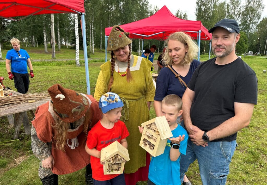 Markku Tiihonen (oik.) ja Sini Puonti sekä pojat Otto (alhaalla oik.) ja Eero Tiihonen tarkastelevat rakentamiaan hyönteishotelleja metsätonttujen Varpun ja Naavan (vas.) kanssa.