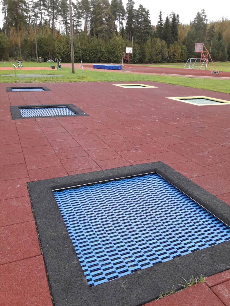 Trampoliinipuisto Ypäjällä Pertunkaaressa. Sininen maahan upotettu trampoliini erottuu punertavasta pinnasta. Trampoliineja näkyy useita urheilukentän reunalla.