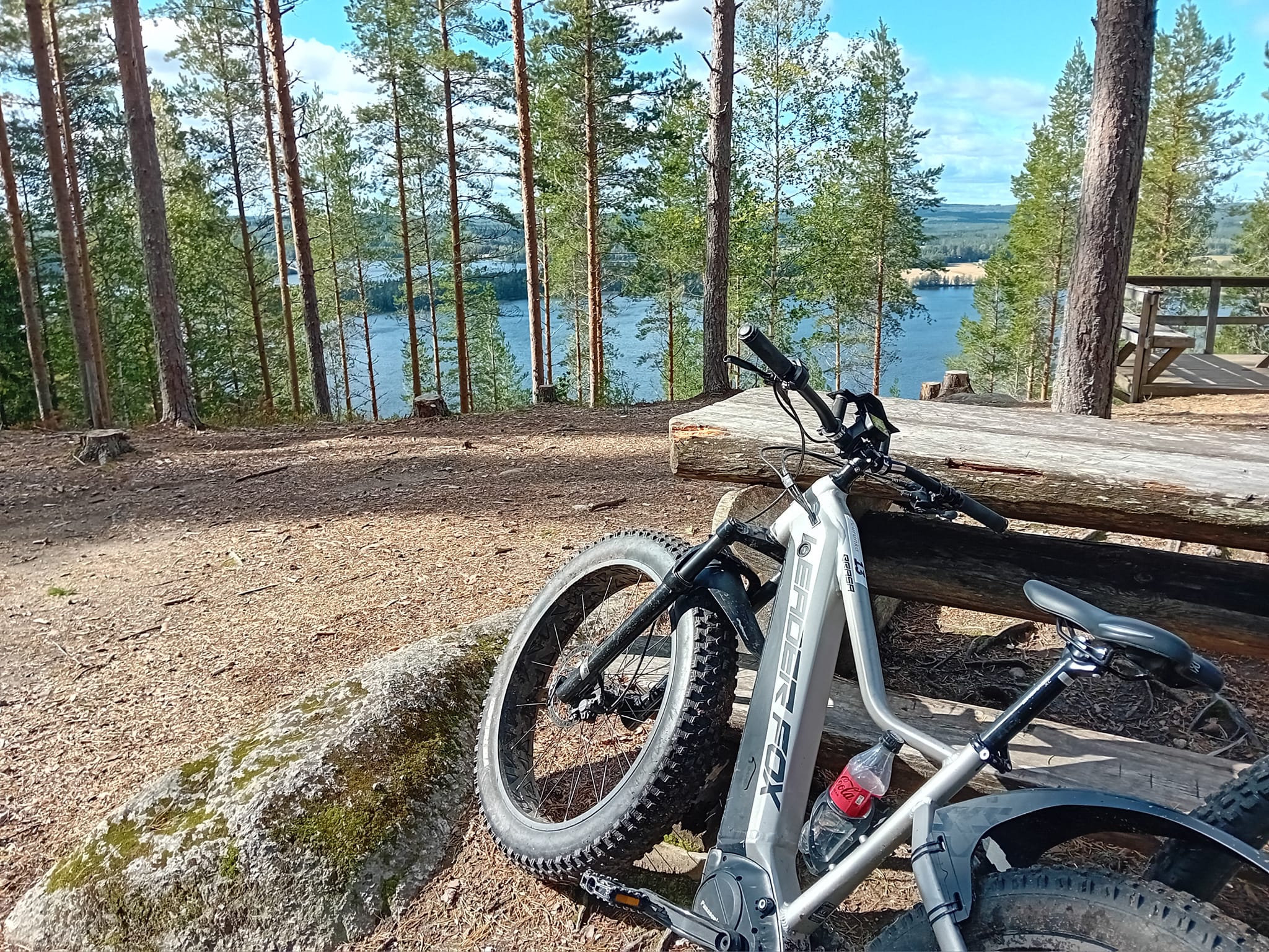Maastopyörä nojaa puiseen pöytään ulkoilureitin pysähdyspaikalla metsässä, taustalla siintää järvi. Paikkakunta Laukaa.