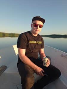 Nuori mies veneessä tyynellä järvellä, kädessä oluttölkki ja päässä lippalakki. 