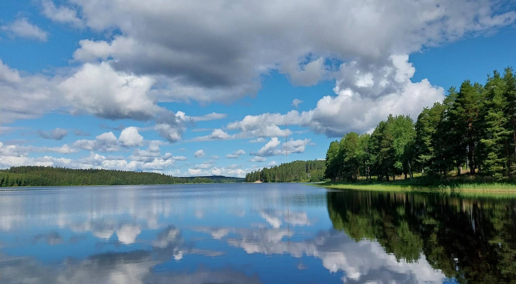 Järvimaisema kesäpäivänä, sininen taivas, kumpupilvet heijastuvat järven veteen.