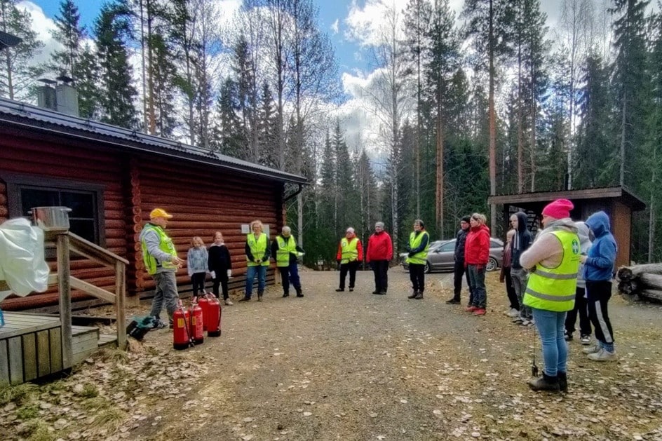 Joukko ihmisiä kuuntelemassa puhujaa kylätalon pihalla, muutamilla päällä huomioliivit.