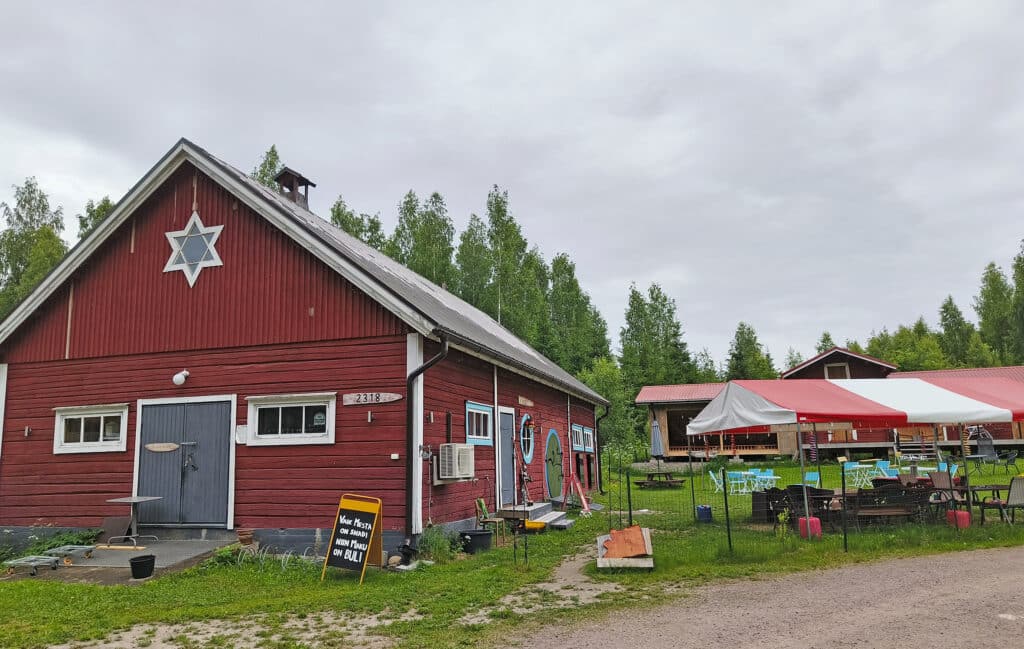Punainen hirsirakennus, jonka päädyssä on iso tähti. Rakennuksen edessä on punavalkoinen telttakatos ja puutarhakalusteite.