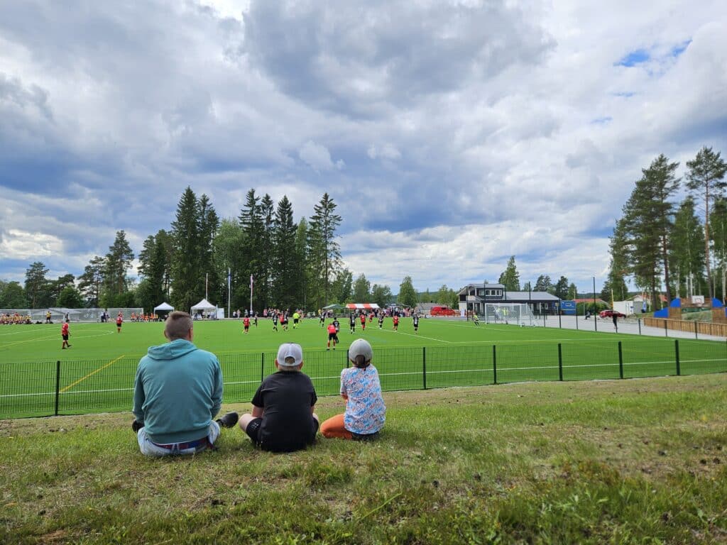 Kolme ihmistä istuu jalkapallokentän laidalla selin kameraan.