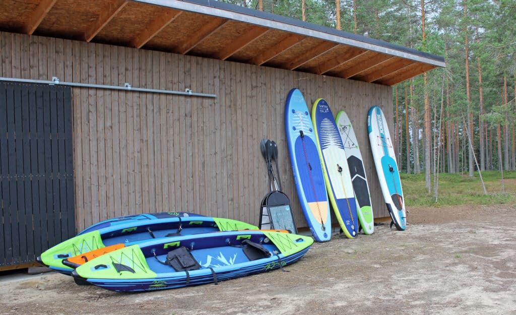 Sup-lautoja nojaa puisen rakennuksen seinää vasten. Maassa on kajakkeja.