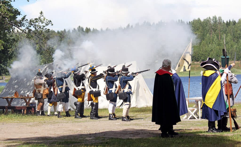 Ryhmä 1700-luvun sotilasasuihin pukeutuneita miehiä seisoo järven rannalla ja tähtää aseilla vastarannalle.