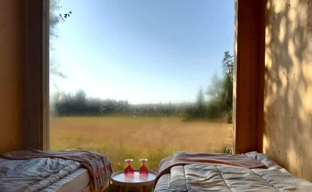 Majoitusmökki sisältä. Kuvassa näkyy ikkunasta avautuva peltomaisema, sängyt ja kaksi punertavaa juomalasia yöpöydällä.