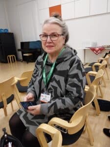 Harmaahiuksinen nainen istuu tuolilla luentosalissa.