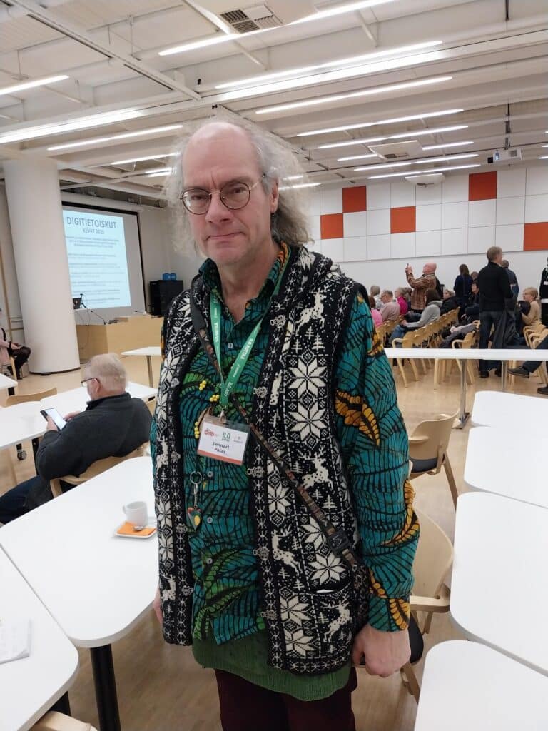 Harmaahiuksinen mies seisoo luentosalissa, tqausalla ihmisiä.