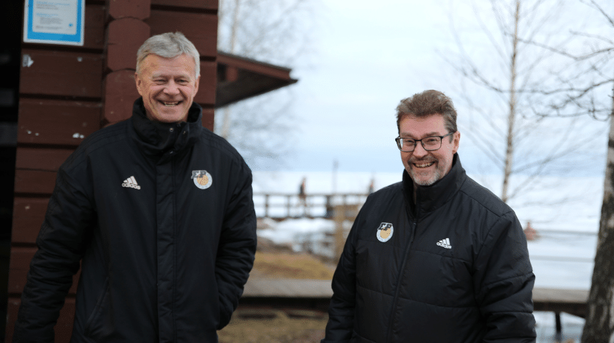 Samanlaiseen verryyttelytakkiin sonnustautuneet miehet nauramassa ulkona rantamaisemassa.