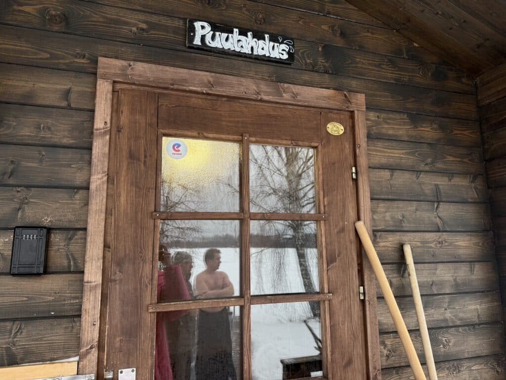 kuva saunan ovesta, teksti Puulahdus, ovesta heijastuu ihmisiä