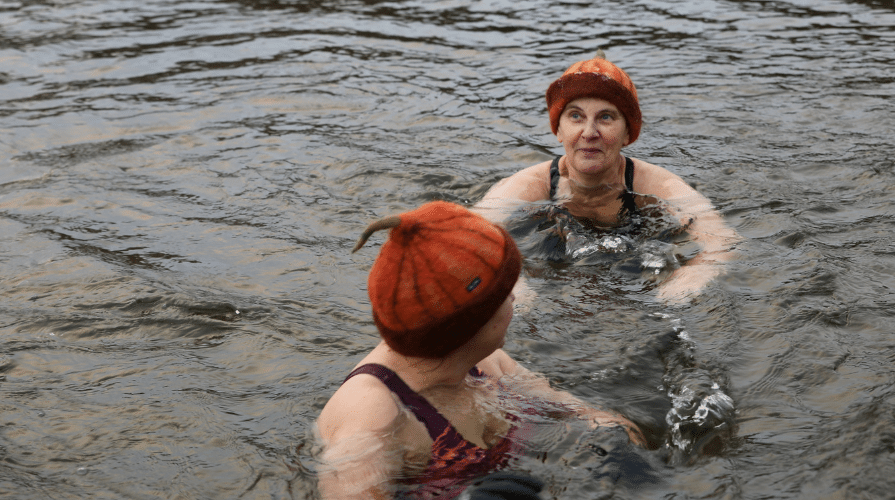 Kaksi naista uimassa avannossa kurpitsaa mukailevat huopahatut päässään.