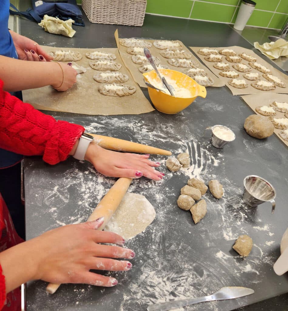 Paistamattomia karjalanpiirakoita pöydällä. Kuvassa näkyy myös kahdet kädet. Toiset kaulitsevat piirakan kuorta toiset rypyttävät.