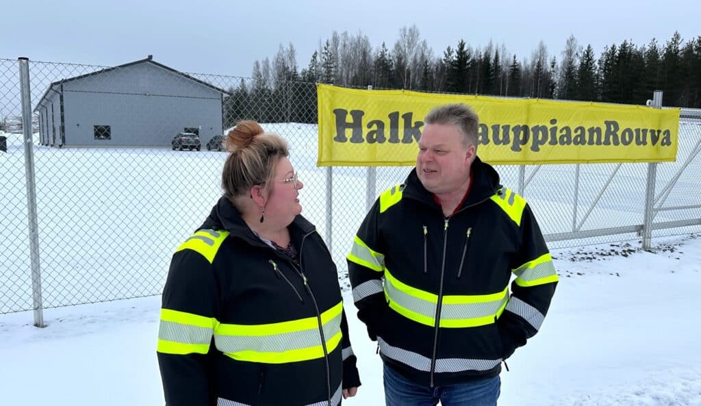 Nainen ja mies hallin ja aidan edessä työvaatteissa. Aidassa mainoslakana "Halkokauppiaan Rouva".