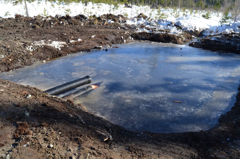 Vettä täynnä oleva maa-allas metässä, altaan reunasta pistää esiin pari muoviputkea.