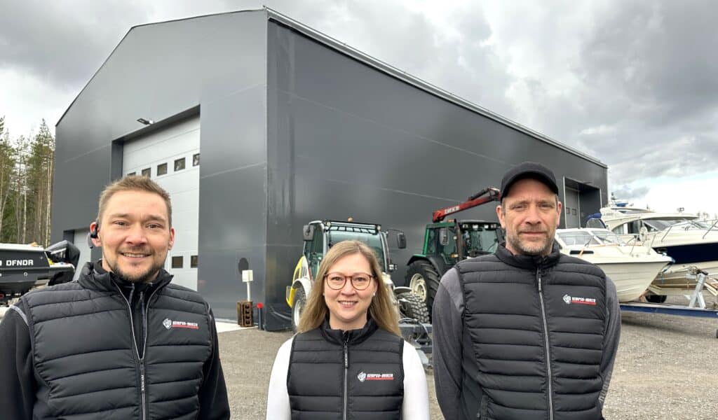 Kolme henkilöä, joiden taustalla harmaa hallirakennus, veneitä ja traktori.