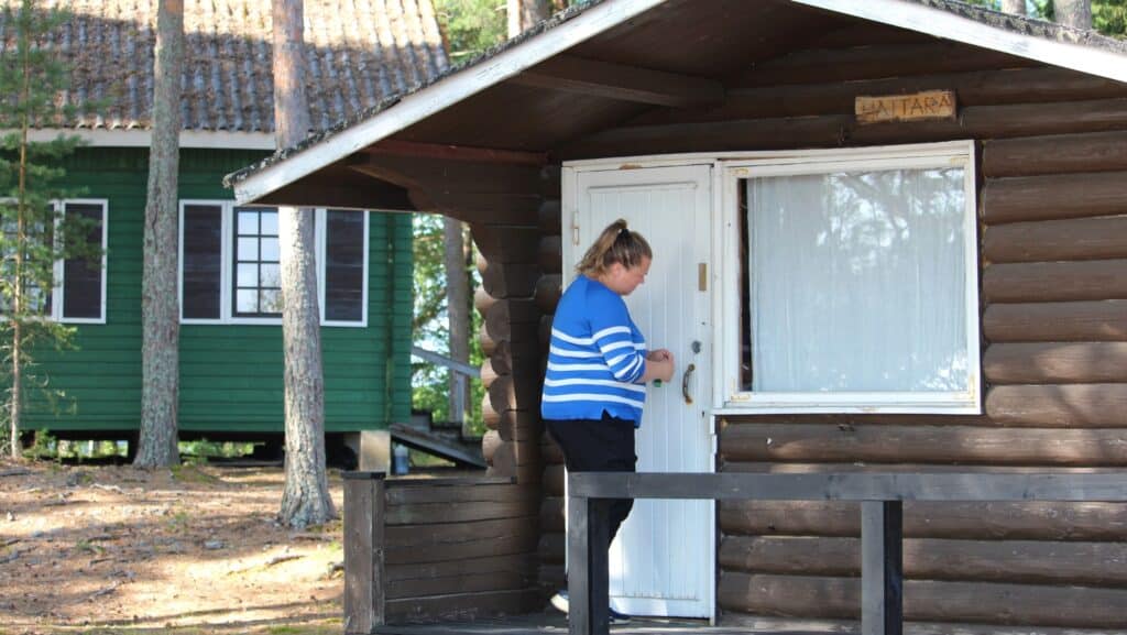 Nainen leirintämökin ovella. Taustalla näkyy isompi puurakennus ja männyn runkoja.