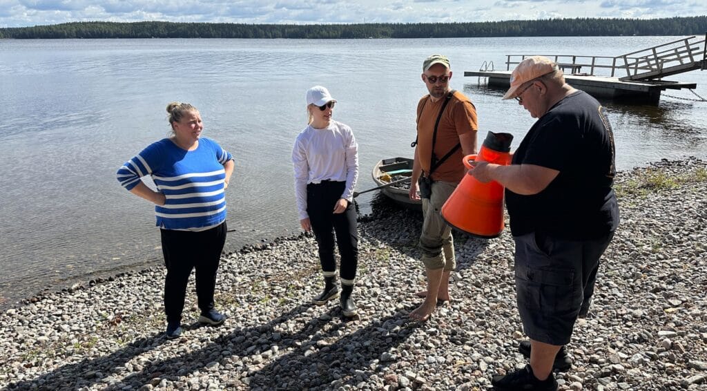 Neljä ihmistä aurinkoisella rannalla. Yksi heistä katsoo oranssia vesikiikaria.