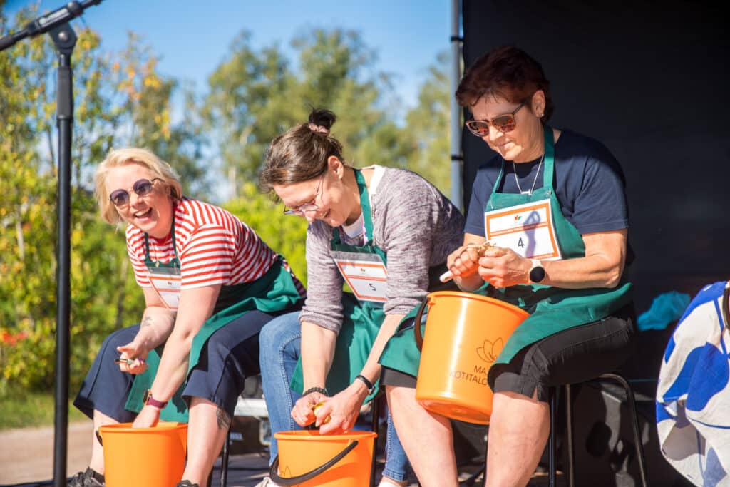 Kolme naista lavalla kuorimassa perunoita.