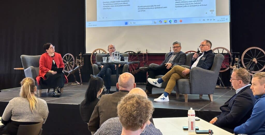 Ihmisiä istumassa nojatuoleissa esiintymislavalla, etualalla yleisöä.