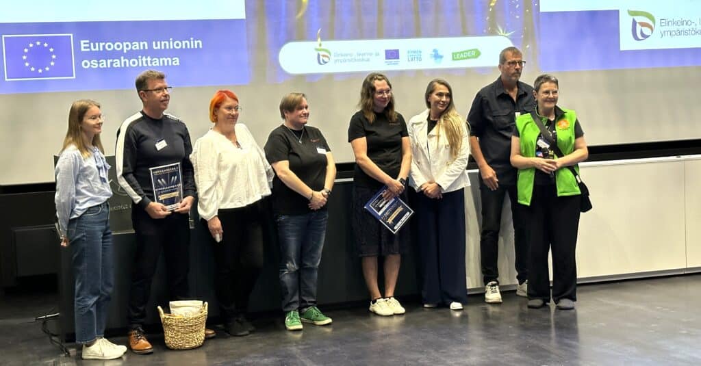 Ihmisiä esiintymislavalla kunniakirjojen kanssa. Taustalla näkyy logoja, esim. Euroopan unionin osarahoittama.