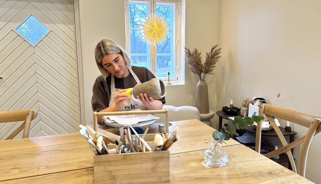 Nainen muotoilee pöydän ääressä saviruukkua. Taustalla ikkunassa palaa joulutähti. Etualalla työkalupakissa pensseleitä ja muita työvälineitä.