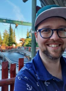 Lähikuvassa hymyilevä nuori mies, jolla silmälasit ja lippalakki.