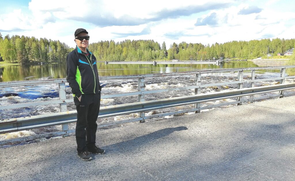 Hymyilevä mies aurinkolasit silmillä ja kädet takin taskuissa seisoo asfalttitiellä, taustalla koski.