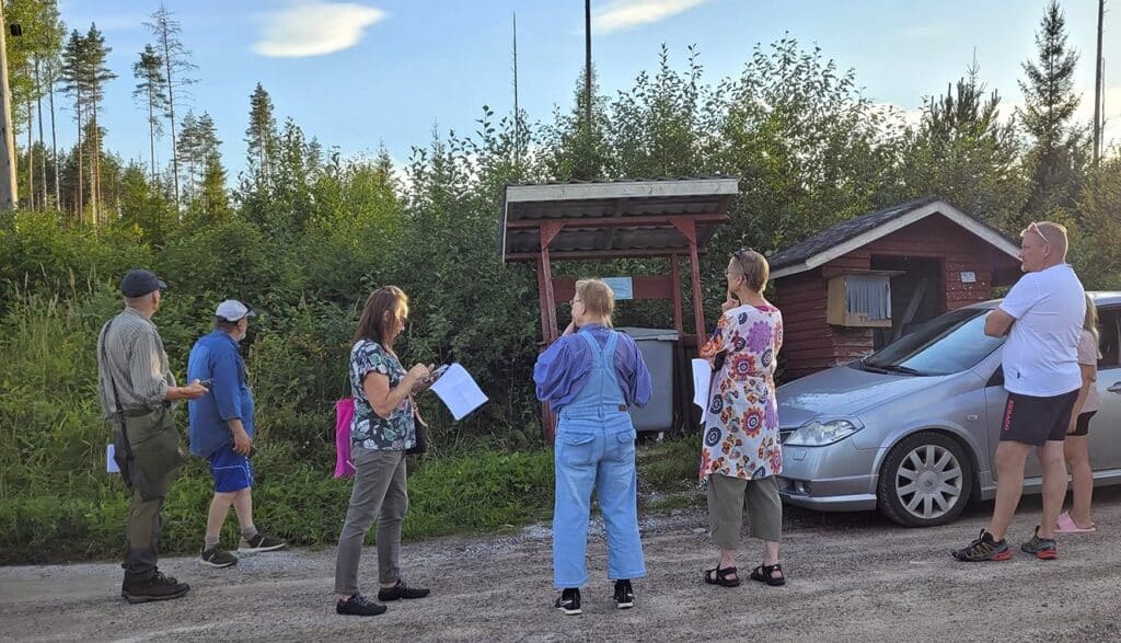 Ihmisiä ja auto maitolaiturin ja opastaulun edessä.