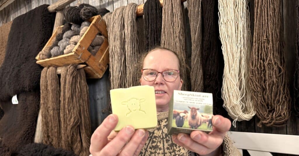 Nainen esittelee saippuaa ja ihovoidepakettia. Taustalla luonnollisen värisiä villalankavyyhtejä.