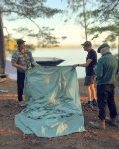 Kolme miestä pystyttää telttaa kesäiltana järven rannalla.