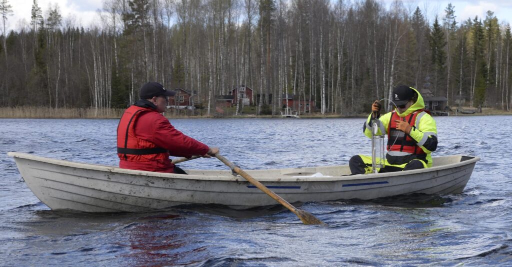 Kaksi miestä soutuveneessä keväisellä järvellä, rannassa puita.
